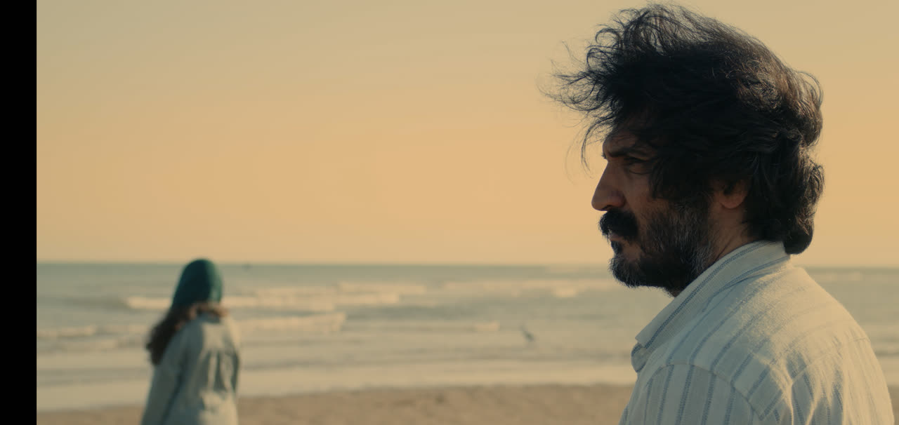 A man with wind-blown hair looks out to sea as a woman walks on the shore