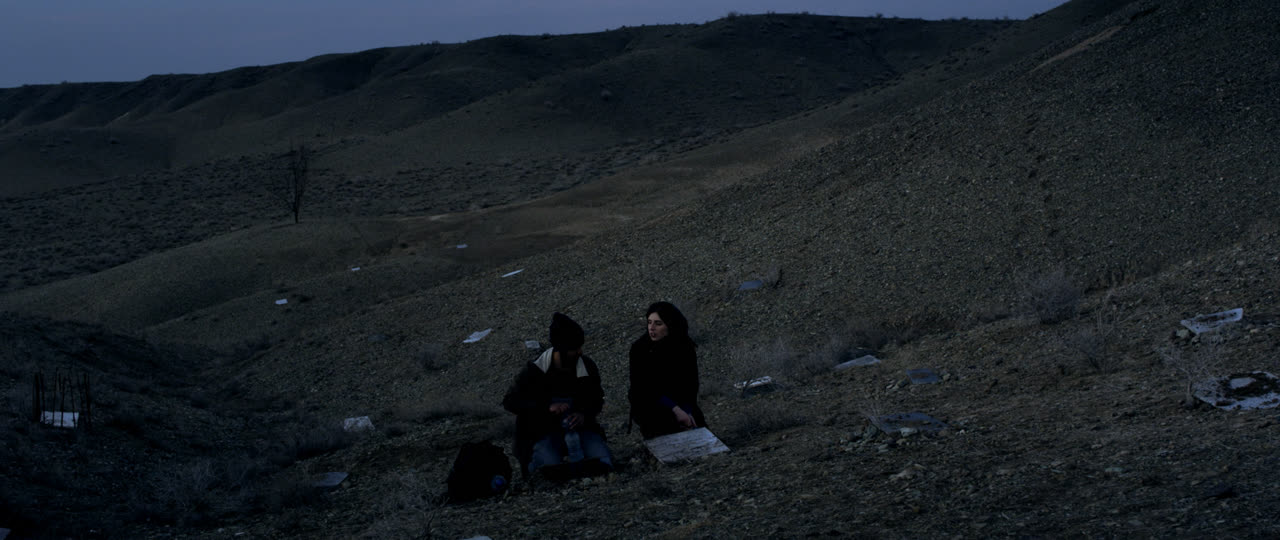Two figures sit on a bare hillside at dusk