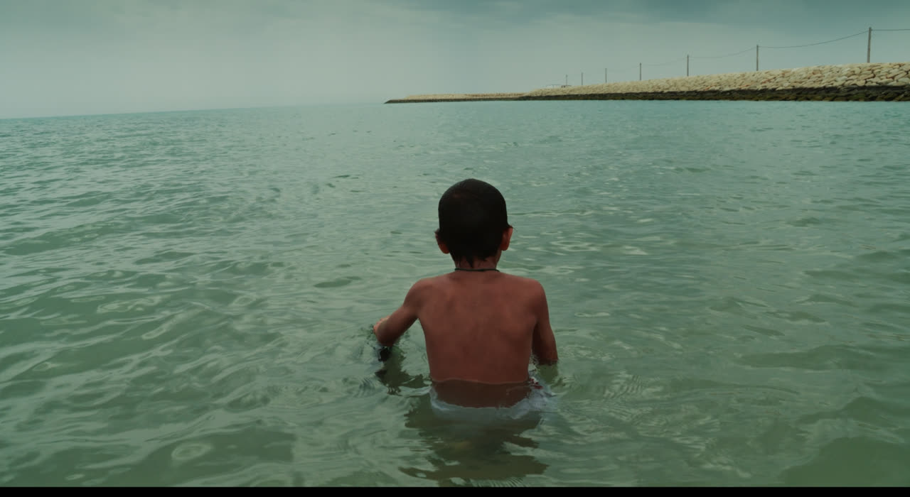 A boy wades into calm sea water beside a long pier