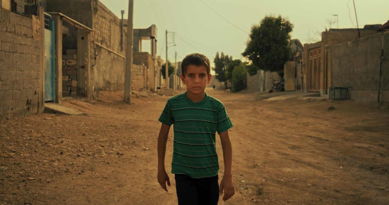 A boy in a green shirt walks down a dusty village street