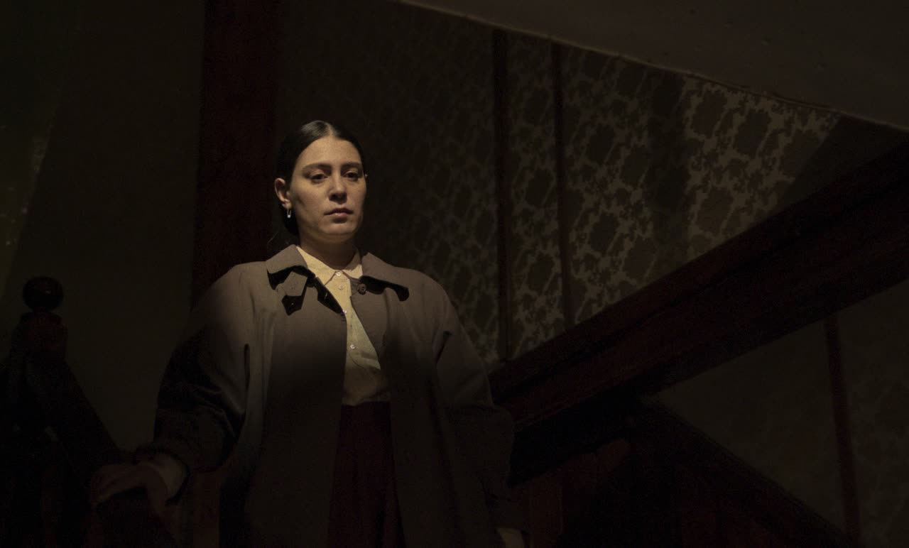 A woman in a trench coat stands at the foot of a dark staircase