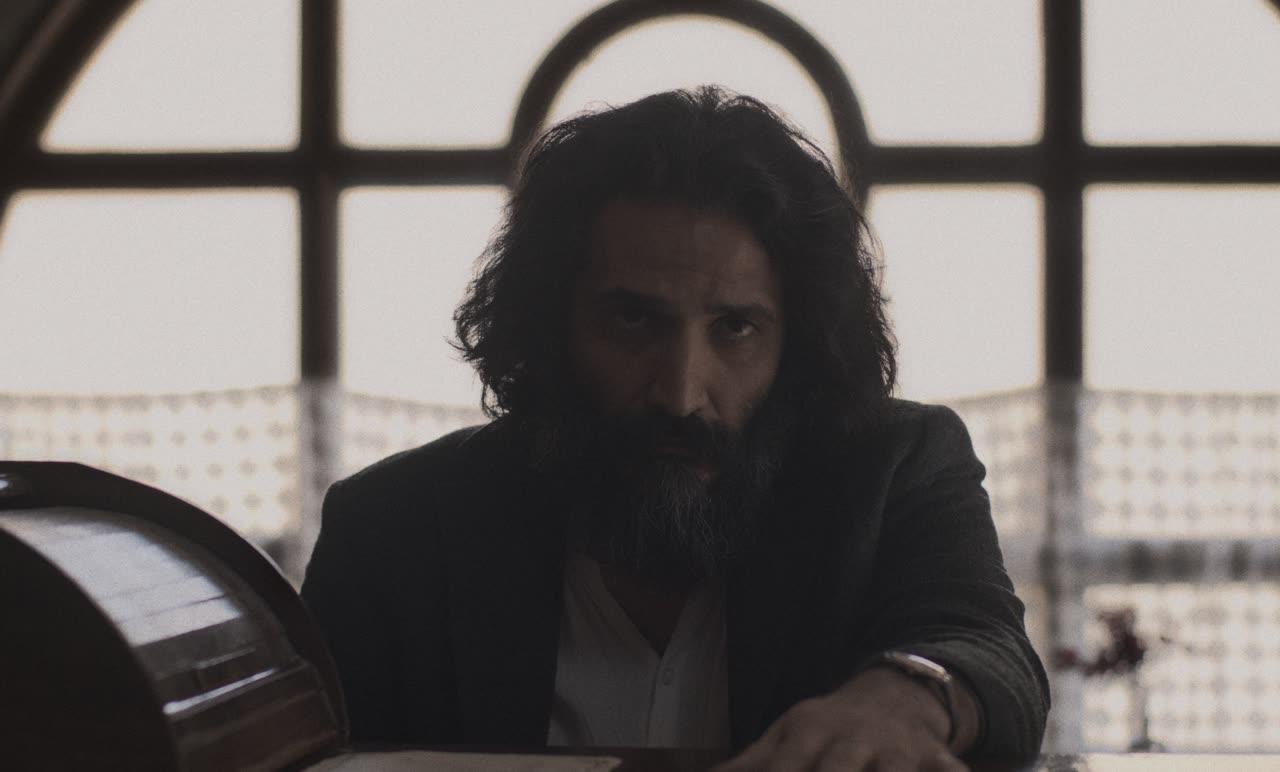 A long-haired man sits alone by an arched window