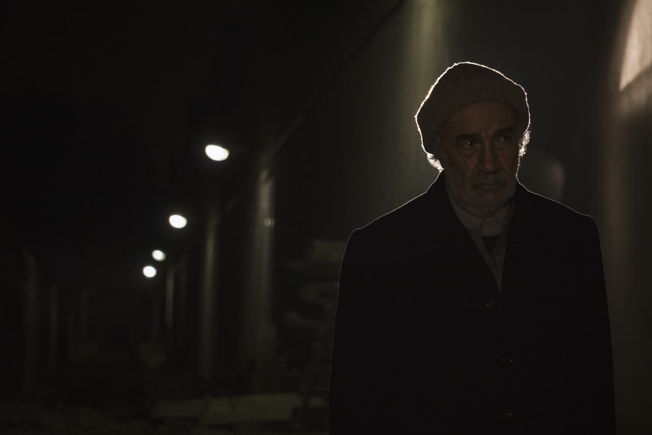 An old man in a dark corridor lit by a row of lamps