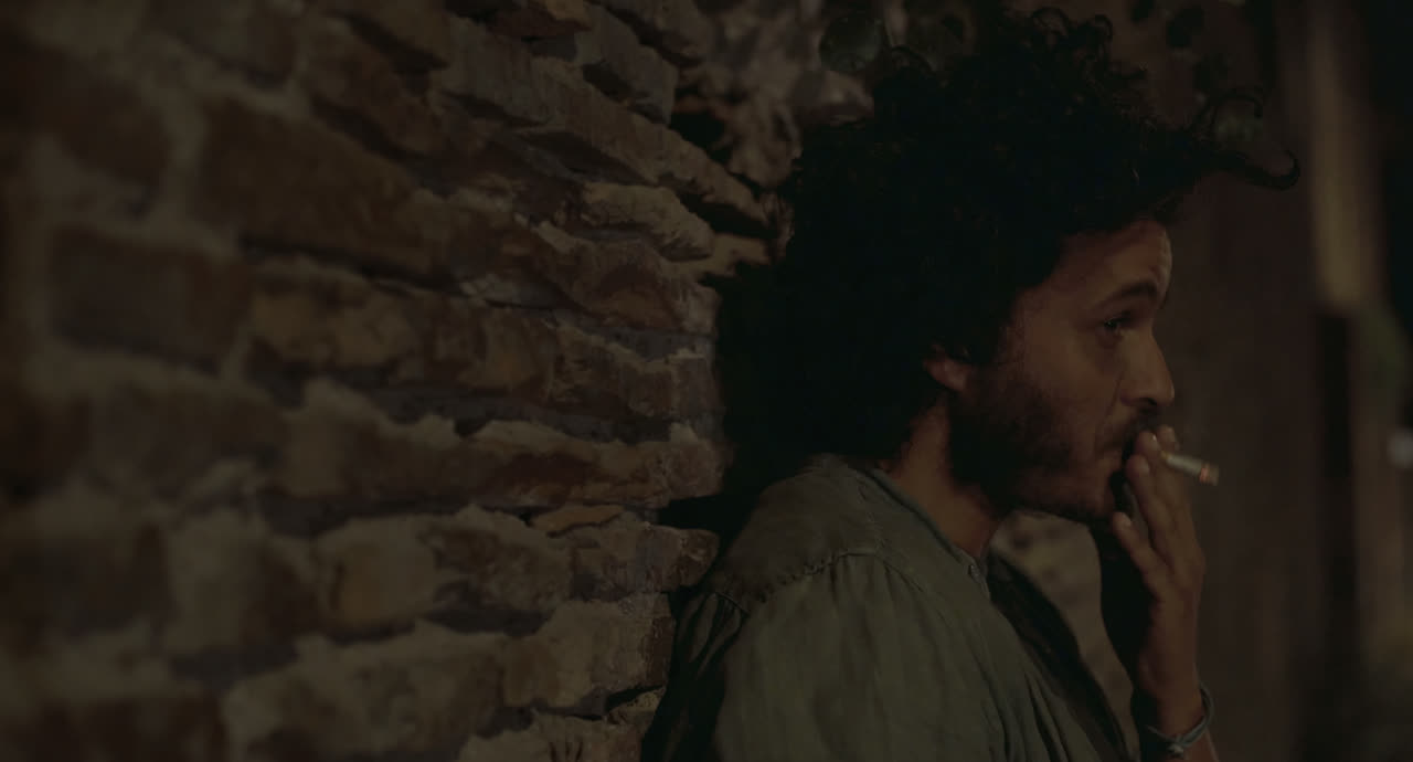A young man smokes against an old brick wall