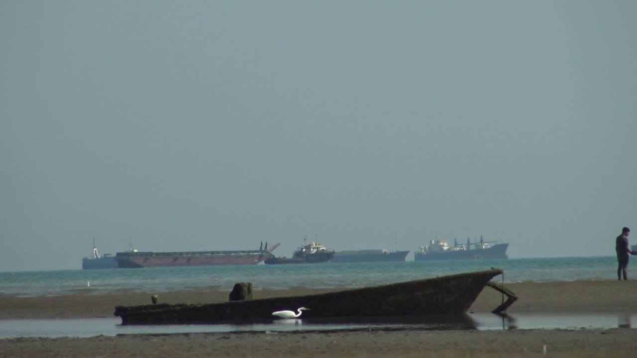 A boat lies on a flat shore with ships on the horizon